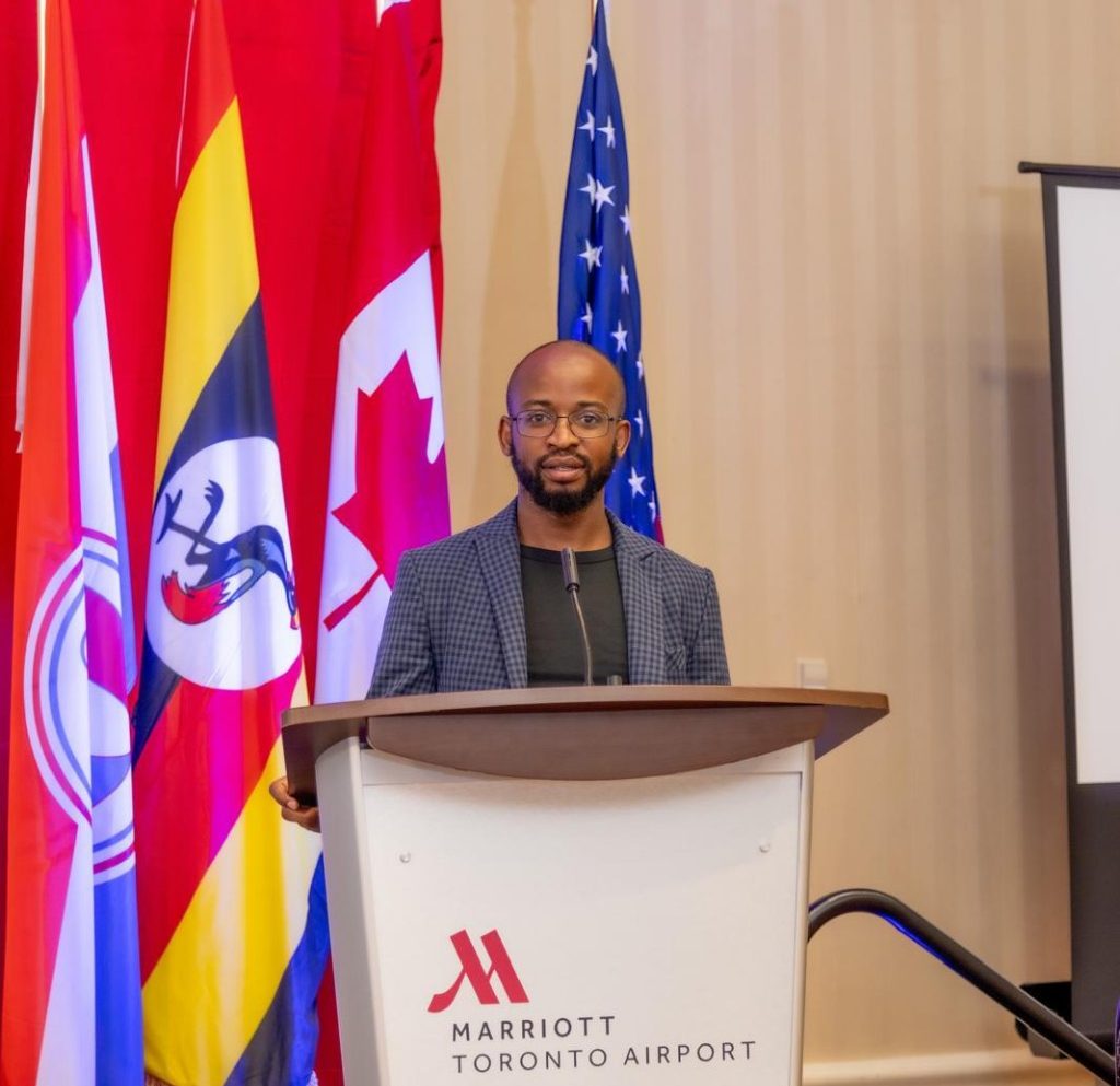 Clinton Mirrors Ampaire, Director of Resurgence Network for Human Rights, stands at a podium during a speech at the Marriott Toronto Airport. Behind him, flags of Uganda, Canada, the United States, and other symbols are displayed, highlighting the international focus of the event. Ampaire is addressing the urgent need for international action against human rights abuses in Uganda under President Museveni's government.