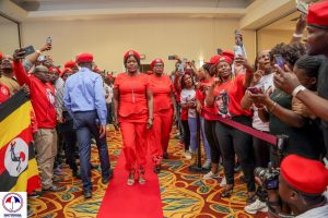 Brenda Nakato, a prominent leader of the NUP Canada chapter, walks confidently down a red carpet flanked by enthusiastic supporters. The crowd, adorned in red NUP attire and holding Ugandan flags, cheers her on while capturing the moment with their phones. Brenda, dressed in a striking red outfit, is the focal point of the scene, embodying the strength and unity of the NUP movement. Her determined stride and the attention of the supporters underscore her role as a key figure in the ongoing struggle for democracy and justice in Uganda. The atmosphere is electric, filled with hope and solidarity as the NUP supporters rally together in a powerful display of collective action.