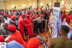 In the image, Brenda Nakato, a prominent leader of the NUP Canada chapter, is seen seated at a table alongside President Robert Kyagulanyi Ssentamu. The two, surrounded by enthusiastic supporters, are engaged in taking pictures, capturing a moment of unity and solidarity. Brenda, dressed in a striking red outfit, stands out as a key figure, reflecting the strong bond between the NUP leadership in Uganda and the diaspora community in Canada. Supporters, many of whom are wearing NUP's signature red berets and shirts, gather around the table, holding up phones to snap photos with the President. The atmosphere is filled with excitement and camaraderie, showcasing the deep connection and shared commitment to the movement's goals. This image represents not just a gathering of supporters, but a powerful statement of collective action and unwavering support for the NUP's mission to bring about change in Uganda