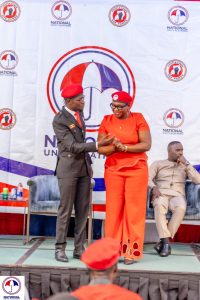 In the image, President Robert Kyagulanyi Ssentamu is seen sharing a warm moment with Doreen Kajumba, a key leader in the NUP Canada chapter. Both are dressed in their symbolic red attire and matching berets, representing their dedication to the National Unity Platform's mission. The two are engaged in a light-hearted exchange, with Doreen smiling as the President gestures towards her, likely in recognition of her efforts and leadership within the NUP diaspora community.