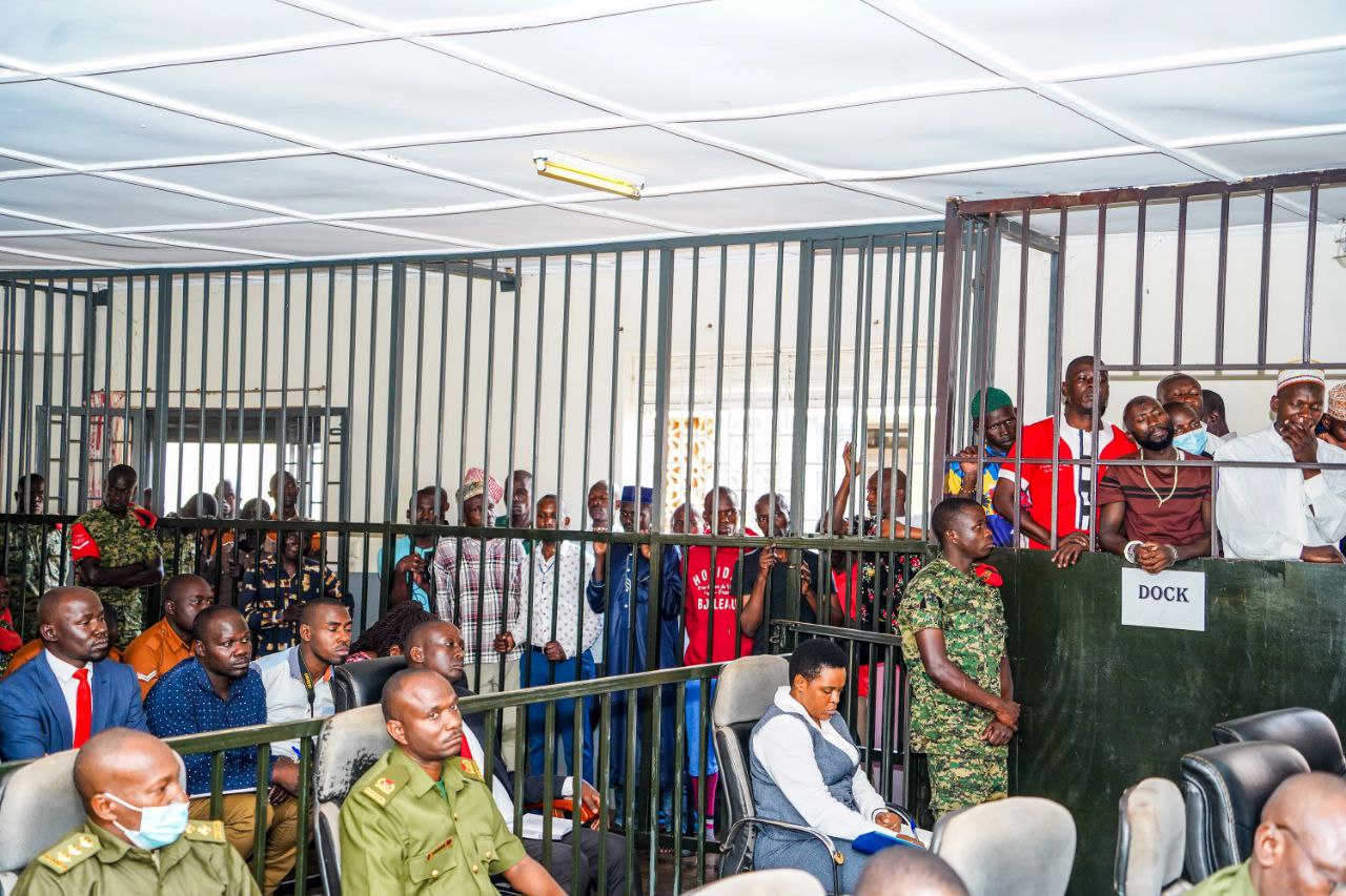 A group of detainees, including Olivia Lutaaya, standing in a courtroom before a panel of military judges. The tension is palpable as they await the decision on their plea bargains after years of legal battles. Some detainees appear distressed, reflecting the pressure they have faced to plead guilty to charges brought against them. Olivia stands tall, her expression determined, symbolizing her resilience despite the state's coercion. The courtroom is filled with military personnel, lawyers, and family members, emphasizing the weight of the case and its political implications.